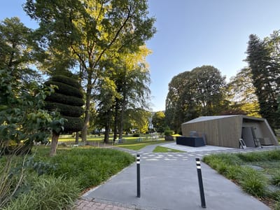 This media file depicts an outdoor scene in a park setting, likely in Bad Orb, Germany. The image is dominated by lush greenery, with large trees providing shade and a clear blue sky overhead, suggesting it is daytime, possibly late afternoon or early evening due to the warm, directional sunlight illuminating the foliage.

In the foreground, a paved walkway leads into the park, flanked by vibrant green grass and ornamental plants. Two black bollards with white reflective stripes are positioned along the walkway, serving as visual markers or barriers.

Further into the park, several white metal chairs and tables are scattered across a manicured lawn, indicating a recreational area or a space for outdoor dining or gatherings. In the middle ground, a uniquely designed wooden building with an angular roof stands out. It appears to be a modern structure, possibly a pavilion or utility building, with its exterior clad in vertical wooden planks. Next to the building, a black covered object, possibly a long table or bench, is visible.

The background reveals more trees and the suggestion of buildings in the distance, hinting at the park's integration with a town or resort. A few indistinct figures can be seen walking in the distance, suggesting a tranquil, leisurely atmosphere. The overall impression is one of a well-maintained public space designed for relaxation and enjoyment of nature. No visible text is present in the image.