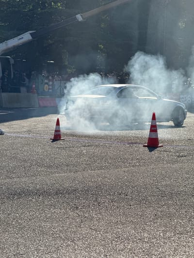 A white coupe is performing a burnout on a paved surface, creating a large cloud of white smoke. Two orange traffic cones with white stripes and the letters "GDS" are visible in the foreground, positioned on the left and right sides of the frame, marking the performance area. In the background, a crowd of people is gathered behind barriers, suggesting an event or competition. A construction crane with the word "NICHI" printed on its boom extends from the top left of the frame, overlooking the scene. The environment appears to be an outdoor location, possibly a street or a dedicated track, with trees visible in the distance. The presence of the crane and barriers indicates that this may be a planned event with spectators. The image does not provide specific details about the city of Poti, Georgia, beyond the context provided in the prompt.