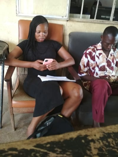 In Bisichi, Nigeria, a woman with braided hair sits on a patterned chair, holding a pink smartphone and a document. She wears a black dress. Beside her, a man in a patterned shirt and maroon trousers sits on a dark couch, looking down. A dark bag is on the floor near the woman's feet. The environment appears to be an indoor waiting area with light-colored walls and a barred window in the background.