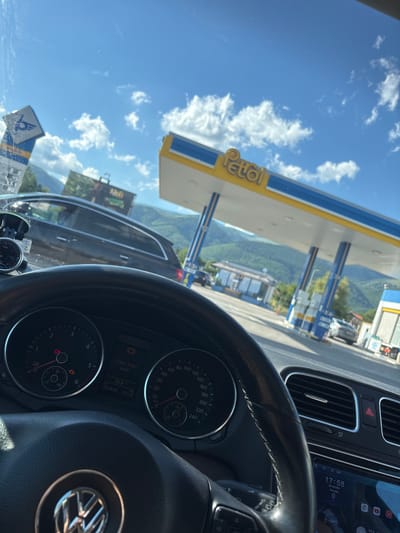 The image shows a view from inside a car, looking out at a Petrol gas station. The gas station canopy is yellow and blue, with "Petrol" clearly visible in yellow letters on the side. In the background, there are lush green mountains under a bright blue sky with scattered white clouds. The scene suggests a sunny day, likely in the afternoon given the clock in the car dashboard shows 17:58.

Inside the car, the dashboard is visible, showing a clock and various indicators. The time is 17:58 on Thursday, June 18, 2026. The temperature is 25.0°C. There's a warning message on the dashboard that reads "Check brake pads!". The steering wheel of the car, with the Volkswagen logo, is also prominent in the foreground. The car appears to be stopped at the gas station, possibly for refueling or a break. The overall impression is one of travel and a brief stop at a roadside service area on a scenic route.