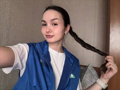 A female individual is depicted indoors, taking a selfie in what is identified as the city of Bayronovka, Russia. She has dark hair pulled back into a long, twisted ponytail, which she grasps with her right hand. Her left arm is extended towards the camera, suggesting she is holding the device. She wears a white short-sleeved t-shirt beneath a blue zippered vest. The vest features Cyrillic text on the left chest, reading "ФОНД ПРЕЗИДЕНТСКИХ ГРАНТОВ" (Presidential Grants Fund), and a green triangular logo on the right. A light-colored smartwatch is visible on her left wrist, and a ring is present on her right ring finger. She has a faint smile and direct eye contact with the camera. The background consists of a textured light brown wall and a partial view of a dark wooden furniture item or door to her right. A white pillow with a dark patterned design is visible behind her, near the wooden object.