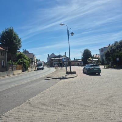 The image depicts a street scene in Bralin, Poland, under a bright blue sky with wispy white clouds, suggesting a sunny day. The perspective is from a slightly elevated position, looking down a paved road.

The road curves to the right, bordered by a cobblestone sidewalk. On the left side of the road, buildings with traditional architecture and some greenery are visible. A dark green car is parked on the sidewalk. Further down the road, a white truck is driving away from the camera. Streetlights are present along the road.

There are several road signs visible, including a blue bus stop sign and various traffic signs indicating regulations or directions. One sign appears to have Polish text, though it's not clearly legible. A traffic light shows a green signal. The overall scene suggests a quiet, possibly residential or small-town commercial area. The sunlight casts distinct shadows, indicating it is likely daytime. No people are prominently visible in the image. The presence of Polish signs confirms the location context.