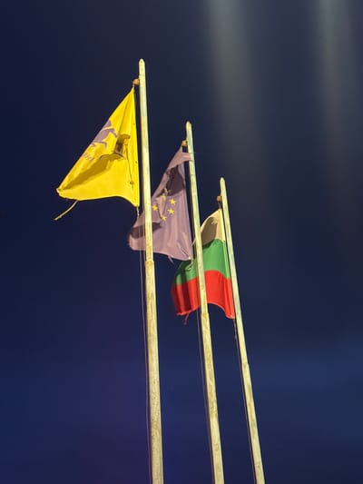 Three flags are displayed on poles against a dark blue background, possibly at night. From top to bottom, the flags are the Bulgarian flag (white, green, red), the European Union flag (blue with gold stars), and an unidentified yellow flag with dark markings. The scene is located in Glojene, Bulgaria.