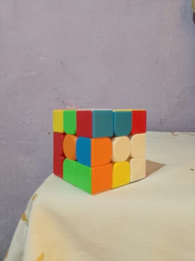 A 3x3 Rubik's cube is positioned on a light-colored tablecloth with a faint yellow floral pattern. The cube is partially solved, with several faces showing mixed colors. The background is a textured, light purple wall. The scene appears to be indoors, possibly a room in Jizzakh, Uzbekistan, given the provided context.
