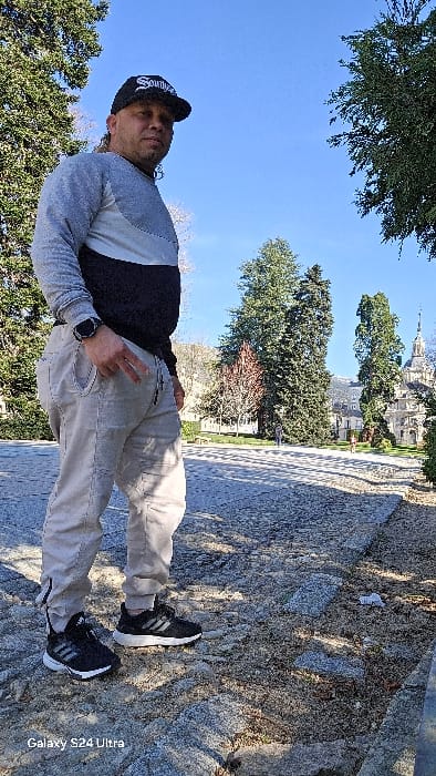 The image depicts an outdoor scene in Real Sitio de San Ildefonso, Spain, on a clear, sunny day. The primary subject is a male, standing on a rough-textured, possibly cobblestone, pathway in the foreground. He is angled to the left, with his head turned towards the viewer. He wears a black baseball cap with white lettering, a long-sleeved crewneck shirt with horizontal blocks of grey, white, and black, light-colored (beige/khaki) joggers with a dark stripe down the side, and black Adidas athletic shoes with white soles. A dark-colored smartwatch is visible on his left wrist. His left hand is partially in his pocket, with his index and middle fingers extended, forming a V-shape. He has facial hair and a visible dreadlock or ponytail behind his left ear.
The environment behind the subject is a landscaped park area. Several tall evergreen trees and some deciduous trees, one with reddish foliage, are visible in the midground. A paved path traverses the park, where two distant human figures are observed walking away from the camera. In the background, a large, ornate baroque-style building with multiple domes and a prominent central spire, consistent with the Collegiate Church of the Holy Trinity at the Royal Palace of La Granja de San Ildefonso, is visible. The sky is bright and clear blue. Shadows from unseen objects or foliage are cast across the foreground pathway. A "Galaxy S24 Ultra" watermark is present in the lower-left corner of the image.