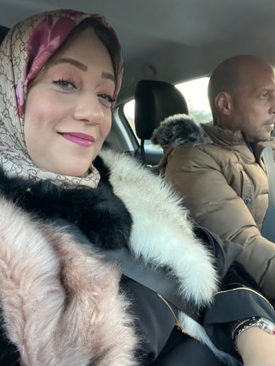 This image captures two people inside a car, seemingly taken as a selfie. In the foreground, a woman is smiling and looking towards the camera. She wears a patterned headscarf, featuring a design resembling the Gucci logo in beige and maroon. Her makeup includes eyeshadow and glossy pink lipstick. She is dressed in a dark coat with prominent fur trims in white, black, and a light pinkish-brown color. A silver-toned watch with a black strap and a fine gold chain are also visible on her.

Behind her, slightly out of focus, a man is seen in the passenger seat. He is bald, looking forward or slightly upwards, away from the camera. He wears a light brown puffer jacket with dark fur trim on the cuffs.

The setting is clearly inside a vehicle, with car seats and headrests visible. Bright natural light streams in from the windows, indicating it is daytime. The background outside the car is blurred but shows signs of outdoor scenery. The overall scene suggests a casual moment during travel, potentially in Majmaa Tolba, Morocco. Both individuals appear relaxed.