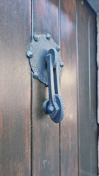 The image shows a close-up, eye-level shot of a dark, ornate door knocker mounted on a wooden door. The door knocker is made of dark metal and features a circular decorative plate with raised elements, from which a hinged arm with a striking plate hangs. The door itself is constructed of vertical wooden planks with a dark stain and visible wood grain. To the right of the door knocker, a light-colored textured surface, possibly stone or a different material, is partially visible. The lighting suggests an overcast day or shade, with soft shadows. The scene is likely located in Rupit, Spain, a town known for its well-preserved medieval architecture.