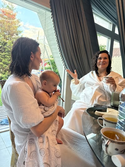 A woman in a white bathrobe holds a baby in her arms, while a second woman in a white bathrobe sits at a table. The baby holds a small wooden spoon in its mouth. The table holds two cups of tea, a container, a tray with a white square of food, and two water bottles. A window behind the seated woman shows a gray curtain and a grid of window panes. A window behind the standing woman reveals green trees, a blue sky, and a white overhang. The location is indoors. The city of Чёрное, Russia, is not directly discernible from the image.