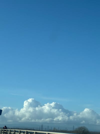 The scene is an exterior view, observed from an elevated position, potentially a bridge or overpass, located in the city of Altenheim, Germany. The upper two-thirds of the frame are dominated by a clear, bright blue sky. A faint, dark, circular artifact is present in the upper-center of the sky. The lower third of the sky features a horizontal band of distinct white cumulus clouds. In the immediate foreground, a light-colored, possibly concrete or painted metal, railing extends horizontally across the bottom edge. A dark, indistinct human figure is partially visible on the far left, positioned adjacent to the railing. Below the cloud line, in the middle ground, a series of dark, slender vertical structures, consistent with industrial pylons or chimneys, are scattered across the distant horizon. To the right, a deciduous tree with bare branches is partially visible. The ambient light indicates a sunny daytime condition.