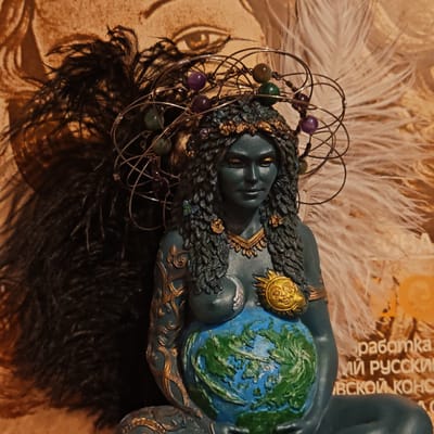 A statue of a blue-skinned female figure with a wire halo and beads sits in front of a backdrop of feathers and blurred text. The figure's belly is painted like the Earth. Her left breast is adorned with a moon and her right with a sun. She wears a crown and a necklace, and her left arm is decorated with gold filigree. The location is Novo-Nikol'skoe, Russia.