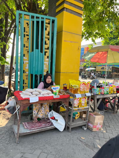 This image shows a street vendor in Juwet Kenongo, Indonesia, selling goods from a wooden cart. The vendor, a woman wearing a black hijab and a pink top, sits behind a red-cloth-covered table laden with white-bagged merchandise, possibly rice. A sign on the table reads "16.500". Adjacent to her cart, a yellow pillar and a turquoise and yellow gate stand prominently. On a tiered shelf to her right, various yellow and red packaged goods, including cooking oil, are displayed, along with a sign indicating "13.000". A colorful umbrella shades some of the merchandise. In the background, other vendors, motorbikes, and people are visible, suggesting a bustling market environment. The ground is paved with grey cobblestones, and trees with green foliage provide shade overhead. The scene depicts a typical street market setting in a populated urban area.