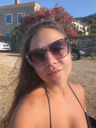 The media file shows a close-up, tilted shot of a young woman lying down outdoors. She is wearing sunglasses with a tortoiseshell frame and a black bikini top. Her face is turned towards the camera, and she appears to be looking upwards or slightly to the side. Her lips are slightly parted, and her expression is relaxed. Her long, brown hair is spread out around her.

In the background, a stone building with blue shutters is visible, partially covered by flowering pink and white bushes. A car is parked behind the building. The ground is covered with dry grass or hay, suggesting a warm, sunny day, likely in the afternoon given the strong shadows. The sky is a clear blue. The scene evokes a sense of vacation or leisure, consistent with the location context of Buljarica, Montenegro, known for its coastal beauty.

The sunglasses have "TOM FORD" visible on the frame. A license plate on the car appears to have some text, but it is not clearly legible. The overall mood is sunny and relaxed.