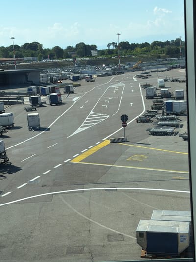 The image shows an aerial view of an airport tarmac in Ferno, Italy. The tarmac is paved with asphalt and marked with various white and yellow lines indicating lanes and directions. Several types of cargo containers and baggage dollies are visible, some loaded onto trailers, suggesting active cargo handling operations. In the background, there are buildings, trees, and a clear blue sky with scattered clouds. The overall scene depicts a functional airport environment with logistical operations underway.