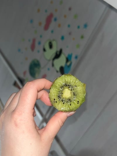 A human hand holds a bisected kiwi fruit. The hand, with fair skin and visible fingernails, grasps the fruit between the thumb and fingers. The kiwi displays a vibrant green pericarp, a lighter yellowish-white central core, and numerous small, black, ovoid seeds arranged radially. The fruit's edges exhibit minor irregularities. The background is out of focus, featuring a light-toned surface adorned with blurred, colorful graphics. These graphics include a stylized green and black cartoon-like figure, possibly holding an object, along with diffuse pink, orange, blue, and green circular and amorphous shapes. The lighting is subdued. The scene is located indoors in Kozlovka, Russia.