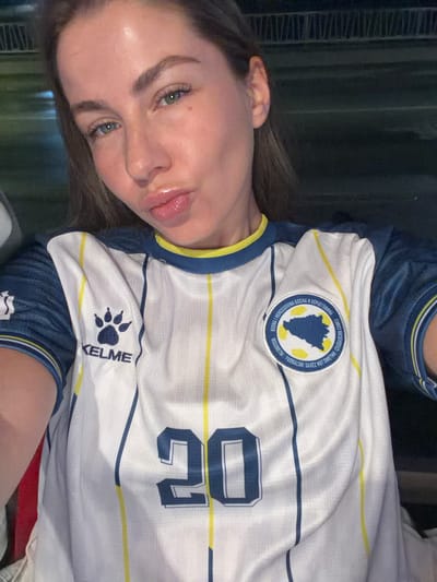 The image shows a close-up, selfie-style photograph of a young woman wearing a white and blue soccer jersey with yellow trim. The jersey has the number "20" prominently displayed on the front. A circular crest with a map of Bosnia and Herzegovina is visible on the left side of the jersey, along with text in Cyrillic and Latin scripts. The woman has her lips puckered, and her eyes are looking towards the camera. Her hair is brown and pulled back. The background is dark and out of focus, suggesting an indoor or nighttime setting, possibly a stadium or a dimly lit room. There are no discernible features in the background that would indicate a specific location within the city of Travnik, Bosnia and Herzegovina.