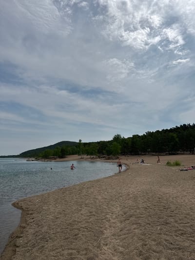 The image depicts a sandy beach adjacent to a body of water, with a treeline forming the background. Several people are visible on the beach and in the water. On the sand, some individuals are sunbathing or relaxing on towels, while others are standing or walking. In the water, a few people are swimming or wading. The water appears calm, and the sky is partly cloudy. The environment suggests a natural recreational area. The location is identified as Ayyrtau, Kazakhstan.