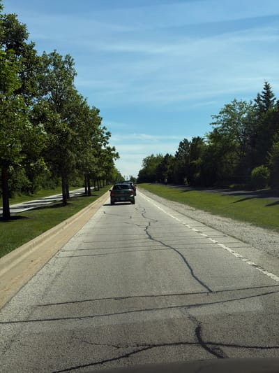 This is a vertical, eye-level shot of a road in Vernon Hills, Illinois, USA, on a clear, sunny day with a light blue sky dotted with faint clouds. The road is paved with asphalt and shows signs of wear, including visible cracks and painted white lines. On the left side of the road, a manicured lawn is bordered by a concrete curb. Beyond the lawn, a line of mature deciduous trees with lush green foliage runs along the roadside. On the right side of the road, a similar line of trees, including some evergreens, borders a grassy area.
Two vehicles are visible on the road, both traveling away from the camera. The closest vehicle is a dark green SUV, possibly a Mini Countryman, with a license plate that reads "AT 94Z9". Behind it, another vehicle, appearing to be a dark sedan or hatchback, is partially visible. The sunlight casts shadows from the trees onto the road and the grassy areas. The overall scene depicts a peaceful drive through a suburban or park-like environment.