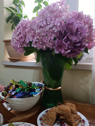 In the city of Adzina, Abkhazia, a domestic scene unfolds with a still life composition on a wooden table. Dominating the background is a large bouquet of pink hydrangeas in a ribbed green glass vase with a gold band. To the left, a white ceramic bowl overflows with individually wrapped chocolates. In the foreground, a white plate holds several slices of layered cake topped with crumbs. The setting suggests a comfortable indoor environment, possibly a dining area or living room, with a window visible in the background.