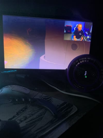 The image captures a desk setup in a dimly lit room, possibly a home office, with a computer monitor as the central focus. The monitor displays a video feed of a man sitting in a chair, likely a streamer or content creator, with a backdrop of electronic equipment. Below the monitor, a purple fan with a digital display showing "2" and a green power indicator is visible. A computer mouse with a patterned mousepad rests in the foreground. The overall setting suggests a gaming or work environment. The image is taken from a slightly elevated angle, looking down at the desk. There are no clear indicators of the specific location within Glodeni, Moldova, or any specific city.
