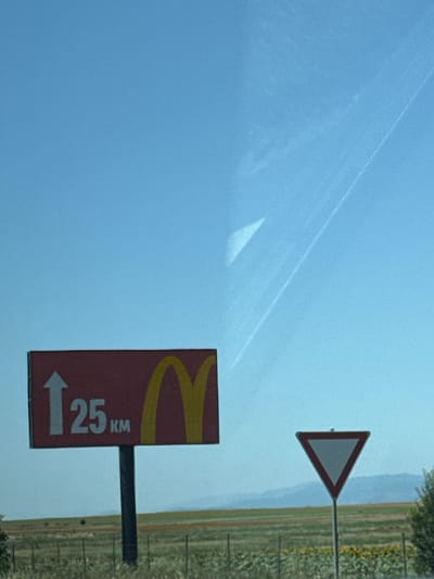 The image shows a roadside scene with a clear blue sky. In the foreground, there is a red sign with a white arrow pointing upwards and the text "25 KM" in white, alongside a yellow McDonald's logo. To the right of this sign, a triangular "Give Way" sign is visible. The background consists of a green field with some trees and distant hills under a bright sky. The image appears to be taken in Vodenichane, Bulgaria, based on the context provided.