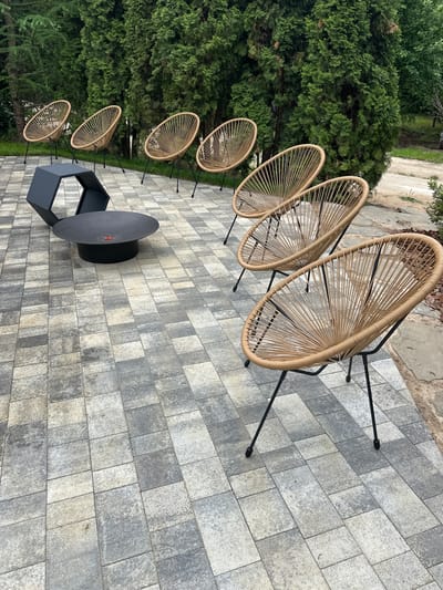 In an outdoor setting, likely a garden or patio area in Zakharovo, Russia, a collection of modern outdoor furniture is arranged on a paver patio. The scene features six tan woven chairs with black metal legs, positioned in two rows. In the foreground, three chairs are clustered, while three more are situated behind them. Adjacent to the chairs, a low, circular dark gray fire pit with a red ignition element is visible, along with a dark gray, angular, hexagon-shaped side table. The background is dominated by lush, dense green trees, suggesting a natural or park-like environment. The lighting indicates it is daytime, with soft natural light. There are no people or animals present in the image.