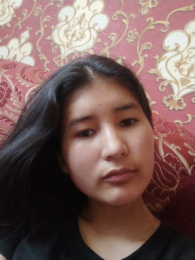 The media file shows a close-up selfie of a young woman with long, dark hair against a background of patterned wallpaper and a cushion. She is looking directly at the camera with a neutral expression. The wallpaper has a red base with ornate gold and cream floral motifs. The cushion is red with a similar embroidered pattern. The lighting suggests it is daytime, possibly indoors. There is no visible text in the image. The location context provided is "Социализм, Kyrgyzstan," which suggests a possible geographic and ideological setting.