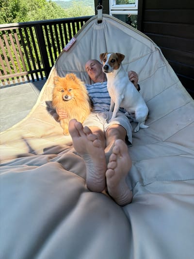 In Lommedalen, Norway, a man is relaxing in a suspended chair on a deck. He is wearing a striped shirt and shorts. Two dogs, a fluffy orange Pomeranian and a white Jack Russell terrier with brown markings, are also in the chair with him. The Pomeranian is to his left, and the Jack Russell is to his right, sitting up. The man's feet are visible in the foreground, facing the camera. The deck overlooks a lush green landscape with trees and a distant hillside. The weather appears sunny and warm, indicated by the shadows and bright lighting.
