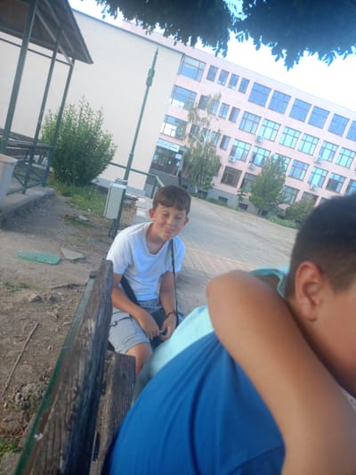 A young boy with brown hair is sitting on a wooden bench, looking to his right with his eyes closed and a slight smile. He is wearing a white t-shirt and grey shorts. A dark strap is visible across his chest, and he is holding a dark object in his right hand. Behind him, another person's arm and shoulder are visible, wearing a blue shirt. The bench is weathered and appears to be outdoors, with a dirt and gravel ground. In the background, a light-colored building with many windows is visible, and there are trees and bushes. The scene appears to be in the city of Mineralni Bani, Bulgaria, likely near a school.