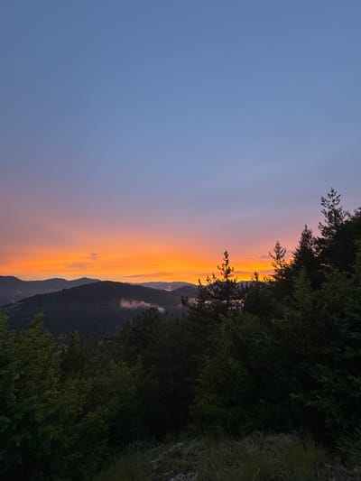 The image depicts a mountainous landscape at dusk. The sky is a gradient of soft blue at the top, transitioning to hues of pink and vibrant orange near the horizon, indicating sunset or sunrise. Layered, dark green mountains recede into the distance, with some patches of white mist clinging to their slopes. In the foreground, a dense forest of evergreen and deciduous trees is visible, silhouetted against the colorful sky. The trees are lush and green, with some taller pine trees reaching upwards. The overall scene is serene and natural, evoking a sense of peace and the beauty of the wilderness. The location is the city of Strazha, Bulgaria.