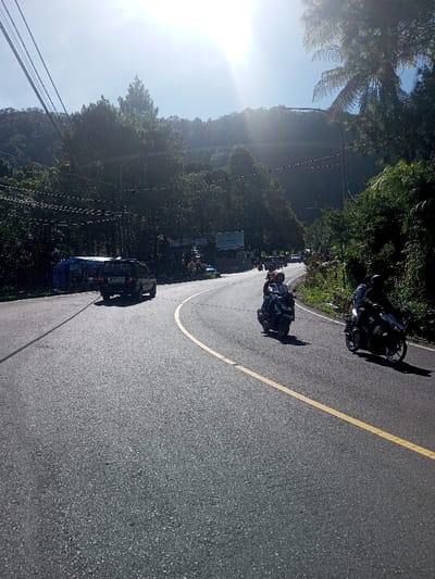 The image captures a curved asphalt road in Ka Thaung (Upper), Myanmar, under bright sunlight, with a prominent sun flare in the upper sky. The road features a continuous yellow line delineating the inner edge of the curve on the right.
Two dark-colored motorcycles are visible on the right side of the road, both being ridden by individuals wearing helmets. The rider of the lead motorcycle wears a light-colored helmet, while the rider of the second motorcycle wears a dark helmet. In the mid-ground on the left, a dark sport utility vehicle is proceeding away from the viewer, displaying a partial license plate of "L.G. 19" on its rear. Additional vehicles, including motorcycles and cars, are visible in the distance along the curving road.
The environment is characterized by dense green foliage and trees lining both sides of the road, ascending into steep, tree-covered hills or mountains that dominate the background. Overhead utility wires are visible in the upper left, supported by a pole. A partially visible blue tarp structure is present on the left roadside, behind the dark vehicle. Multiple billboards with indistinct text and graphics are discernible on the left side of the road in the mid-distance. The sky is clear.