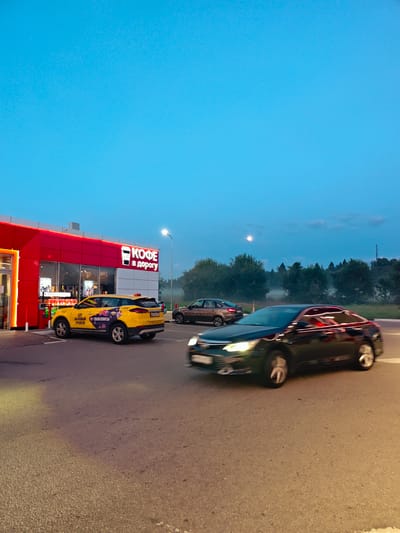 This image captures a scene at dusk outside a red and white building with a sign reading "КОФЕ В ДОРОГУ" (Coffee on the Road), located in Dubrovki, Russia. A yellow taxi is parked to the left of the entrance. To the right, a dark-colored car is in motion, appearing to drive away from the scene. In the background, a line of trees is visible against a pale blue sky with some clouds and a faint moon. The ground is asphalt, suggesting a road or parking area. The lighting indicates it is either early morning or late evening. No people are clearly visible within the frame.