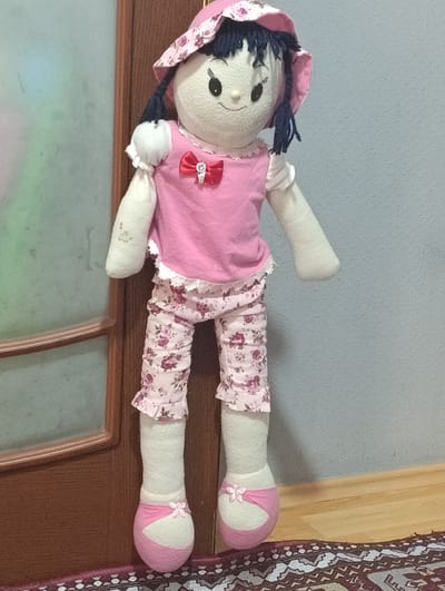 A stuffed doll is standing upright against a wall. The doll has a rounded white fabric head with black button eyes, a small red button nose, and a stitched smile. It has dark blue yarn hair and is wearing a pink hat adorned with a floral pattern. The doll's body is covered in a pink top with white lace trim around the neck and sleeves. The top is decorated with a red bow and a small silver embellishment. The doll is wearing pink pants with a repeating floral pattern of purple flowers. The pants also have white lace trim at the ankles. The doll's arms and legs are made of white fabric. Its feet are covered in pink and white fabric shoes, each decorated with a small pink flower.
The doll is standing next to a wooden doorframe and a wall painted a light blue-grey. The floor is covered with a patterned rug and a wooden baseboard. The lighting suggests it is daytime. The setting appears to be indoors.