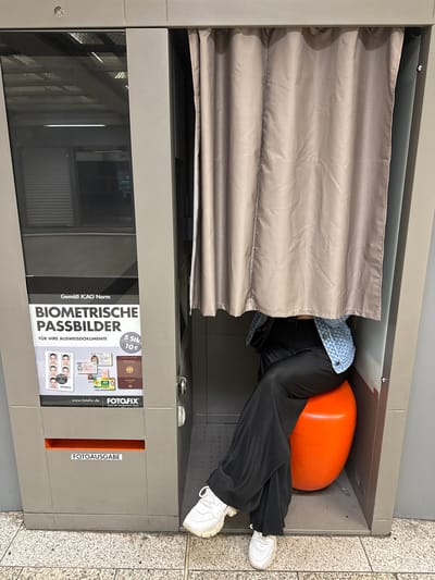 A person is seated in a photo booth, partially obscured by a beige curtain. They are wearing a blue patterned jacket and black wide-leg pants, with white sneakers on their feet. The person is sitting on an orange, egg-shaped stool. To the left of the photo booth is a machine with text in German advertising "Biometrische Passbilder" (Biometric Passport Photos) and pricing information. The floor is tiled with a speckled pattern. The scene appears to be in a public indoor space, possibly a train station or shopping center, based on the lighting and general architecture. This image was taken at the Duisburg Hauptbahnhof (Duisburg Central Station) in Duisburg, Germany.