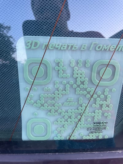 A light blue, 3D-printed sticker is adhered to the inside of a car window. The sticker has the text "3D печать в Гомеле" at the top, which translates to "3D printing in Gomel." Below this text is a QR code, with an Instagram logo stylized within it. Further down, there is a "VOLVO STARGLASS" logo and certification information: "DOT 497 M-41 AS2," "E9 43R-000470," "FRANCE," and "CCC A048587 0." The QR code and text are raised from the surface of the sticker. The background shows the interior of a car, with a dark fabric headliner and a hint of the car's exterior visible through the window, including trees and a silhouette of a person. The sticker is positioned on the passenger side window of the vehicle. The scene appears to be outdoors, possibly in sunlight, given the reflections on the glass. The presence of the "Gomel" text on the sticker suggests a potential origin or service location, but the image itself does not provide direct evidence of the sticker's current location in Бабовіцкі сельскі Савет, Belarus.