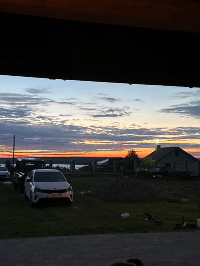 The image depicts a twilight scene, possibly in the village of Пепеляева, Russia, viewed from under a wooden overhang. The sky is filled with dramatic clouds painted in shades of blue, gray, orange, and yellow, indicating the sun is either setting or has recently set. A thin crescent moon is visible in the upper left quadrant of the sky.
In the middle ground, there is a body of water, likely a lake or river, with a distant shoreline dotted with trees and buildings. A dark silhouette of a utility pole with wires stretches across the left side of the frame.
In the foreground and middle ground, several cars are parked on a grassy area. A white Kia Rio with the license plate K007KE 159 is prominently featured. Adjacent to it, other vehicles, including a dark truck-like vehicle, are partially visible.
Further back, there is a residential house with a gray roof and a chimney, illuminated by a bright light on its roof. The area around the house appears to be under some form of construction or landscaping, with piles of dirt and debris. There is also a fence made of concrete posts and horizontal bars. The ground in the immediate foreground is paved with stone tiles.