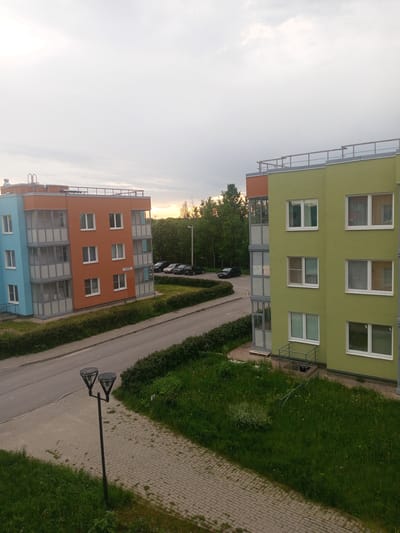 The image displays a residential street scene in Kommunar, Russia, likely during early evening or late afternoon, as indicated by the overcast sky with soft, diffuse light and a hint of warm, low sunlight on the horizon behind the trees.
The primary subjects are two multi-story apartment buildings. On the left, one building is painted in contrasting sections of bright orange and light blue, featuring numerous white-framed windows and several enclosed glass balconies. A white sign on its orange facade clearly shows the Russian text "ул. Енисейская д.11", translating to "Yeniseyskaya Street, Building 11." To the right, a second building, primarily lime green with an orange upper section, also features multiple white-framed windows and balconies.
An asphalt road curves between the buildings, leading to a parking area in the background where several cars are visible. Lush green grass, neatly trimmed hedges, and a line of deciduous trees flank the road and buildings. In the foreground, a paved sidewalk or pathway runs alongside a grassy area, where a distinctive black streetlamp with two conical, upward-facing light fixtures stands tall.
No people are visible, suggesting a quiet moment in the residential area. The scene appears well-maintained and orderly, capturing a typical view of modern housing in the provided location context.
