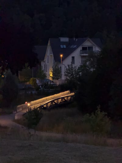 The image depicts a nocturnal scene in the vicinity of Lufingen, Switzerland, focusing on a bridge illuminated by warm-toned lights. The bridge, constructed with wooden elements and a concrete railing, spans a small incline. A circular traffic sign with a red border is affixed to the railing on the left side of the bridge, indicating a prohibition. To the left of the bridge, a paved pathway curves upwards. In the background, a multi-story building with visible windows and a balcony is situated amidst dark trees. A street lamp with an orange light is positioned to the left of the building. The surrounding environment consists of grassy areas and dense foliage, contributing to the overall dim lighting of the scene.