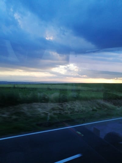 An eye-level, wide shot captures a dusk landscape viewed through a window, likely from a moving vehicle. The foreground is dominated by a dark asphalt road with a bright white line marking its edge, creating a stark contrast against the subdued tones. A blurred, grassy embankment runs alongside the road, suggesting a rural or highway setting. Beyond the embankment, a vast, green field stretches towards a hazy horizon. The sky above is a dramatic mix of deep blues and grays, with scattered white clouds that are illuminated by the fading sunlight, hinting at the approaching night. The overall impression is one of a tranquil, transitional moment in time, with the light suggesting it is either sunrise or sunset.