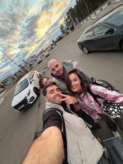 A group of four individuals is captured in a selfie-style photograph taken in Емельяново Airport, Russia. The photo is oriented at an angle, showing a section of the airport parking lot with cars and a sky filled with clouds at sunset. The people are posing close together. One of them is a bald man with a grey hoodie. Beside him is a woman with brown hair, wearing a pink plaid shirt and black pants. Another man to the right is making a peace sign with his fingers, sporting a white jacket. The person taking the photo is presumably a man with visible arm hair in the foreground. The background shows cars lined up in the parking area, including a white car with "taxi" markings. The sky transitions from a bright sunset to a darker evening hue, with golden-colored clouds.