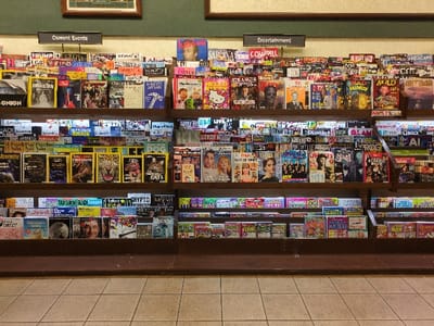 The image displays a well-stocked magazine rack in a retail setting. Two signs at the top read "Current Events" and "Entertainment," categorizing the various publications. Titles cover a wide range of topics, including current events, entertainment, science, nature, and puzzles. Magazines like "The Onion," "Astronomy," "Dinosaurs," "National Geographic Cats," "Taylor Swift," and "Sudoku" are visible among many others, catering to diverse interests.