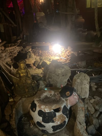 The image captures an outdoor scene in Baan Don Sai, Thailand, at night. A concrete and stone structure houses a collection of decorative items. A weathered, painted duck statue with black and white spots is prominently positioned near the top. Below it, a gold-colored Buddha figure is nestled among the rocks. The overall environment is dimly lit, with a bright light source on the left illuminating nearby foliage and wooden beams. The general impression is one of a grotto-like display with a mix of religious and decorative objects.