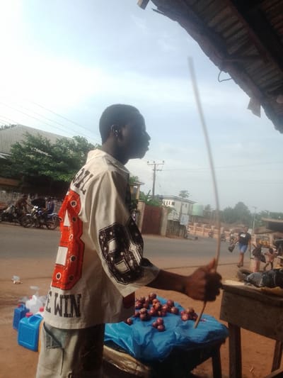 A man stands at a roadside stall selling onions. He wears a white shirt with large numbers and the words "BE WIN" on the back. The onions are piled on a blue tarp. He holds two long, thin sticks in his hand. Behind him, a road stretches into the distance with people, motorcycles, and houses visible. A partial view of a roof is in the upper right corner.