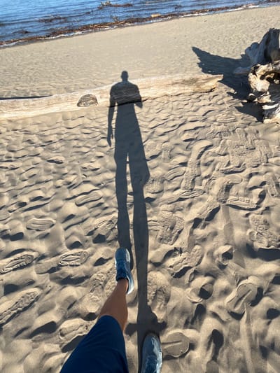 The image depicts a person's legs and shadow on a sandy beach. The person is wearing blue shorts and blue athletic shoes. The sand is textured with numerous footprints and ripples, indicating it is a beach or a sandy area. A large piece of driftwood lies on the sand in the background, and further back, the water of a body of water, likely a lake or sea, is visible, with some debris or vegetation in the water near the shore. The sun is casting a long shadow of the person, suggesting it is either early morning or late afternoon. The location is the shore of the Turka River in Krasnoturansk, Russia.