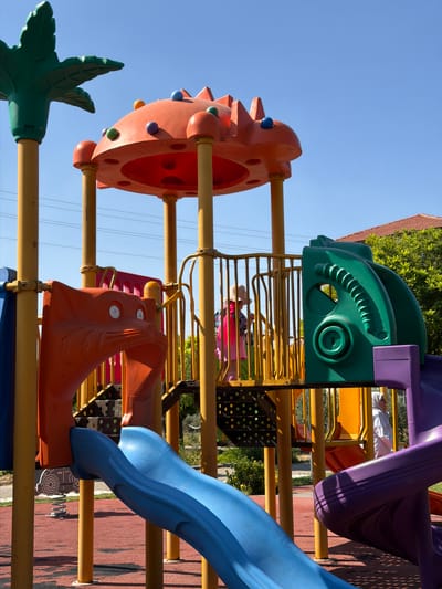 An outdoor, colorful playground structure is visible on a sunny day. The structure is made of various brightly colored plastic and metal components, including slides, platforms, and support poles. A prominent feature is an orange, cat-like structure with wide eyes. A young child wearing a straw hat and a pink dress is on a yellow platform. A second person, wearing a white headscarf and white clothing, is visible in the background. The playground is set on a red rubberized surface, and greenery and a building with a red roof are visible in the background. Electrical wires are strung across the sky. The location is identified as Köprübaşı, Turkey.