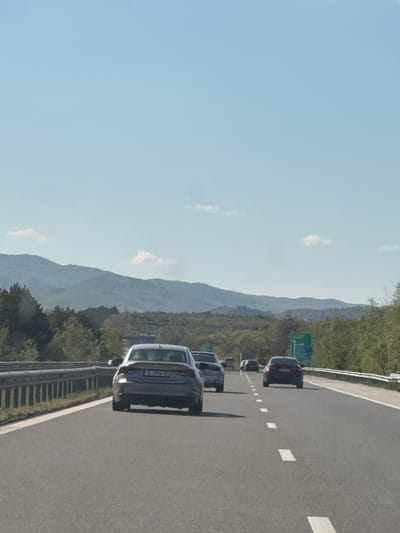 The image, correctly oriented, displays a daytime scene on a highway near Lesichovo, Bulgaria. The sky is bright and mostly clear with a few wispy clouds. The highway itself is an asphalt road with prominent white lane markings, extending into the distance.

Several cars are visible on the highway, forming a queue or moving in moderate traffic. The closest vehicle to the viewer is a dark-colored sedan or hatchback. Its rear license plate, "B 3996 XK", is clearly discernible, indicating a Bulgarian registration from Sofia. Further along the road, more vehicles, predominantly dark-colored, can be seen.

A metal guardrail runs along the right side of the highway, separating it from the adjacent terrain. Beyond the guardrail, a green road sign, partially obscured, is visible. The background features a striking landscape of mountains or large hills, which are densely covered with green trees and foliage, suggesting a natural and possibly rural setting. The overall scene captures a moment of travel on a motorway, framed by mountainous scenery under clear weather conditions. No specific individuals or explicit emotions are visible, but the scene implies ongoing vehicle movement.
