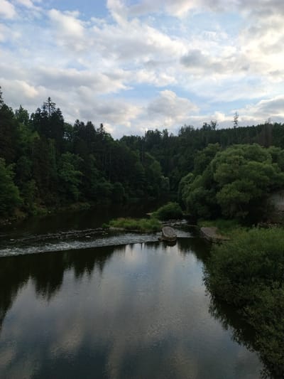 The image depicts a wide, calm river flowing through a dense, green forest. The sky above is partially cloudy with a mix of blue sky and white, gray clouds. The river's surface reflects the surrounding trees and the sky, creating a mirror-like effect. On the right bank of the river, there is a structure made of stone, possibly part of an old building or dam. The river has some ripples and small rapids in the foreground, suggesting a gentle flow. The scene appears to be a natural landscape, likely in a rural or wilderness area. The provided metadata indicates the location is Dobřejice, Czechia.