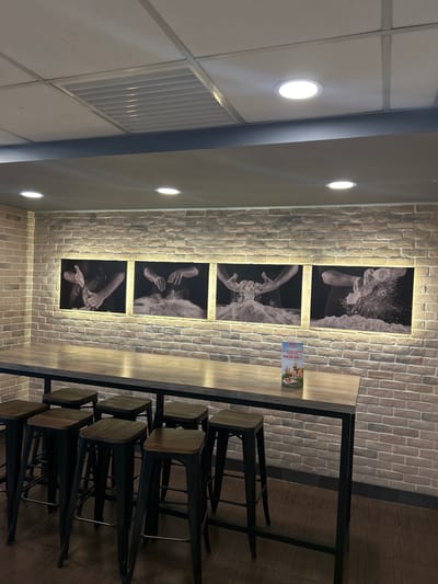 The image presents an interior view of a commercial establishment, likely a cafe or restaurant, in Subang Jaya City Council, Malaysia. The setting features a prominent faux brick-textured wall on the right, illuminated by integrated strip lighting that accentuates its surface.
Decorating this wall are five vertically arranged black and white framed photographs. Each image focuses on human hands engaged in dynamic actions, appearing to be kneading dough, mixing ingredients, or interacting with splashing water, suggesting an artisanal or culinary theme for the venue.
To the right of the brick wall, a long, high-top communal table is visible, featuring a light wooden surface and sturdy black metal legs. Several matching high stools with wooden seats and black metal frames are neatly tucked beneath the table, providing seating for customers.
On the table surface, a small promotional sign displays food images, possibly desserts or ice cream, with the visible text "MY CREAM CHARM COMBO..." and "RM39.90.-".
The ceiling is a grid-patterned drop ceiling, equipped with multiple bright, circular recessed LED lights providing ambient illumination, along with a large rectangular air conditioning vent. The floor is a dark wooden or wood-effect material. No people are present, and the artificial lighting indicates an indoor setting that could be utilized at any time of day. The overall atmosphere appears modern, clean, and functional.