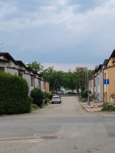 The image depicts a residential street in Hluboká nad Vltavou, Czechia. On the left side of the street, several attached white houses with dark brown roofs and gutters are visible, bordered by dense green hedges. A white car is parked on the street in front of these houses. The right side of the street features more attached houses, one with a yellow facade and a dark roof, another white house, and further down, houses with a light beige facade. A blue and white traffic sign indicating a dead end is posted next to the yellow house. The street is paved with asphalt, and a drainage grate runs along the edge of the sidewalk. In the distance, trees line the street, and several vehicles, including a white camper van and a dark car, are parked further down the road. The sky is cloudy with patches of blue.