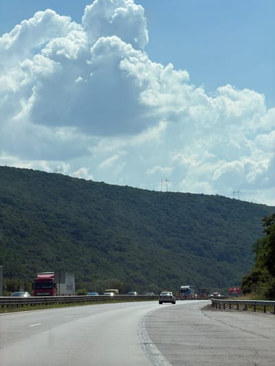 The image depicts a traffic jam on a highway next to a densely forested mountain. The perspective is from a vehicle on the highway, looking ahead and to the left. The sky is a bright blue with large, fluffy white clouds. Two sets of electrical transmission towers are visible on the side of the mountain.
The highway is paved and appears to be curving to the right. A guardrail runs along the left side of the road. A line of vehicles, including cars, trucks, and what appears to be a bus, are stopped or moving slowly. The trucks are large and some have visible signage. One truck has "RURITEX" written on its side. Traffic cones and signs are also present along the side of the road, suggesting potential road work or a detour.
The mountain is covered in lush green trees, indicating it is likely summer or late spring. The sun is shining, and the bright clouds suggest a fair weather day. The traffic jam suggests a potential delay or incident on the highway. The scene is set in Churek, Bulgaria, implying a scenic, mountainous region. No people are clearly discernible in the image. The overall mood is one of a typical travel day with an unexpected delay, set against a beautiful natural backdrop.