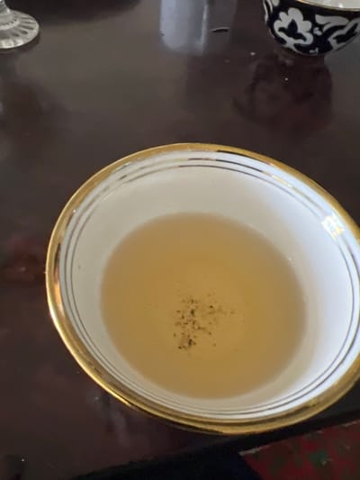 A close-up, high-angle shot shows a white bowl with a gold rim filled with a light-colored liquid, possibly tea. The liquid has a few small dark specks floating on its surface. The bowl is placed on a dark, reflective surface, likely a wooden table, which occupies most of the frame. In the upper right corner, out of focus, is another dark bowl with a white floral pattern. The overall lighting is dim, and the focus is on the bowl of liquid. The image is a still life, with no people or action present. The location is Hazorasp, Uzbekistan.